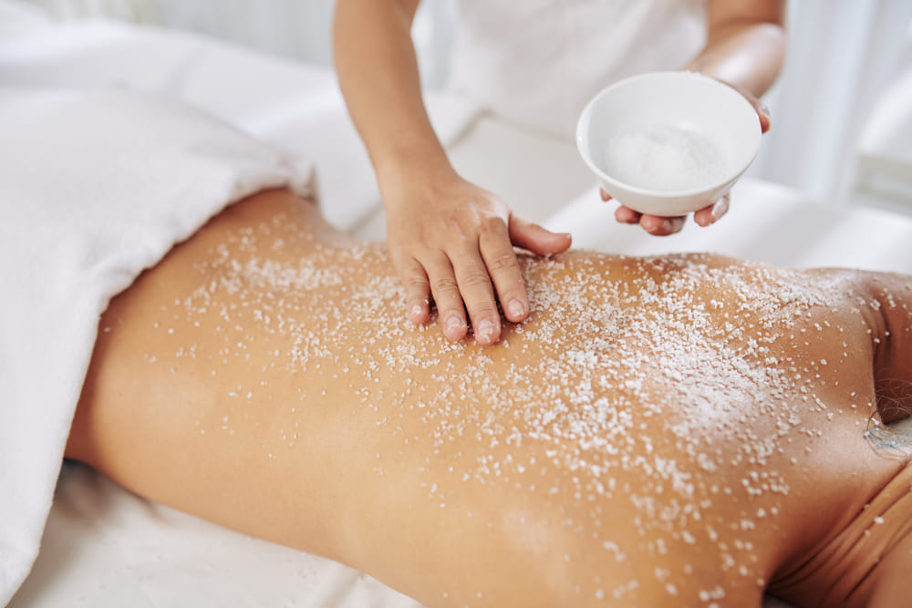 Beautician exfoliating back of young woman with salt scrub before massage