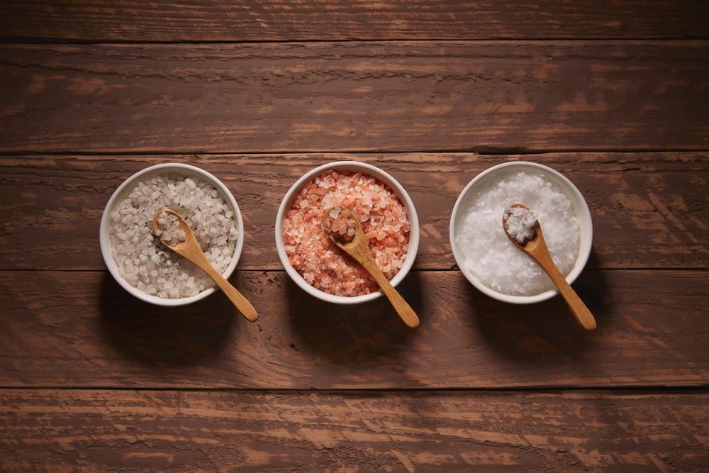 Three ceramic bowls with salt and wooden spoons, the one on the left with salt and herbs, the one in the center with pink salt, the one on the right with flake salt.