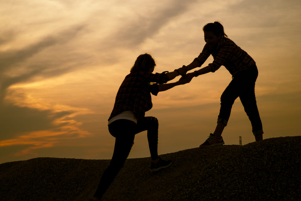Female hikers climbing up mountain cliff and someone giving hand to help partner to success together with sunset background.