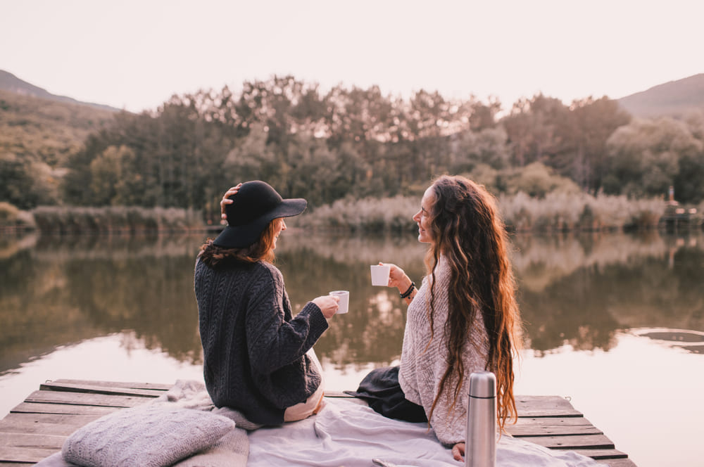 Two female friends in knitted warm sweaters having picnic near lake with autumn forest and lake on the background. Cozy fall atmosphere.