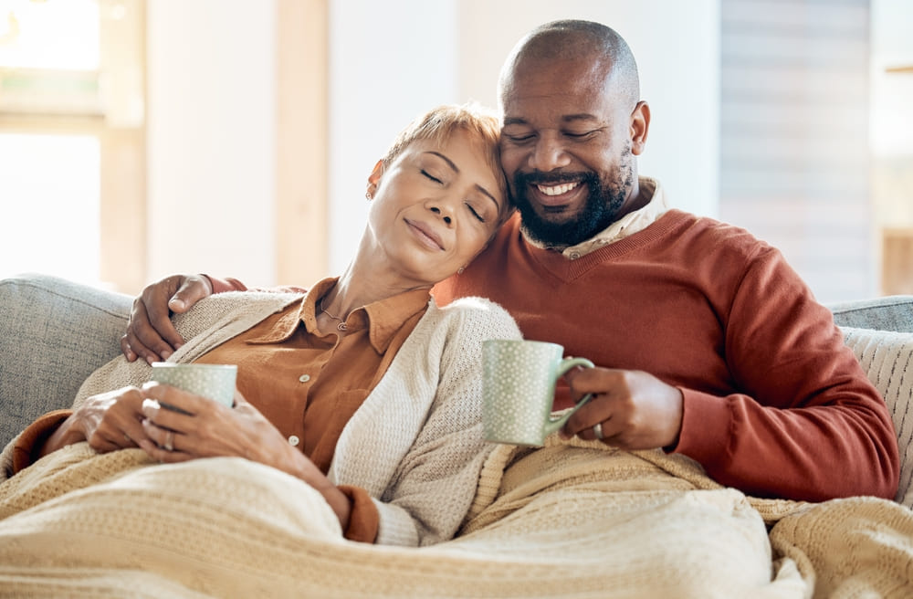 Couple relax on sofa with coffee, blanket and smile on winter weekend morning in home. Peace, comfort and love, happy man and tired woman, cozy time on couch with drink in living room together.