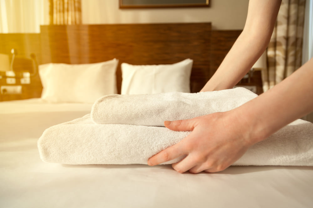 Close-up of hands putting stack of fresh white bath towels on the bed sheet. 